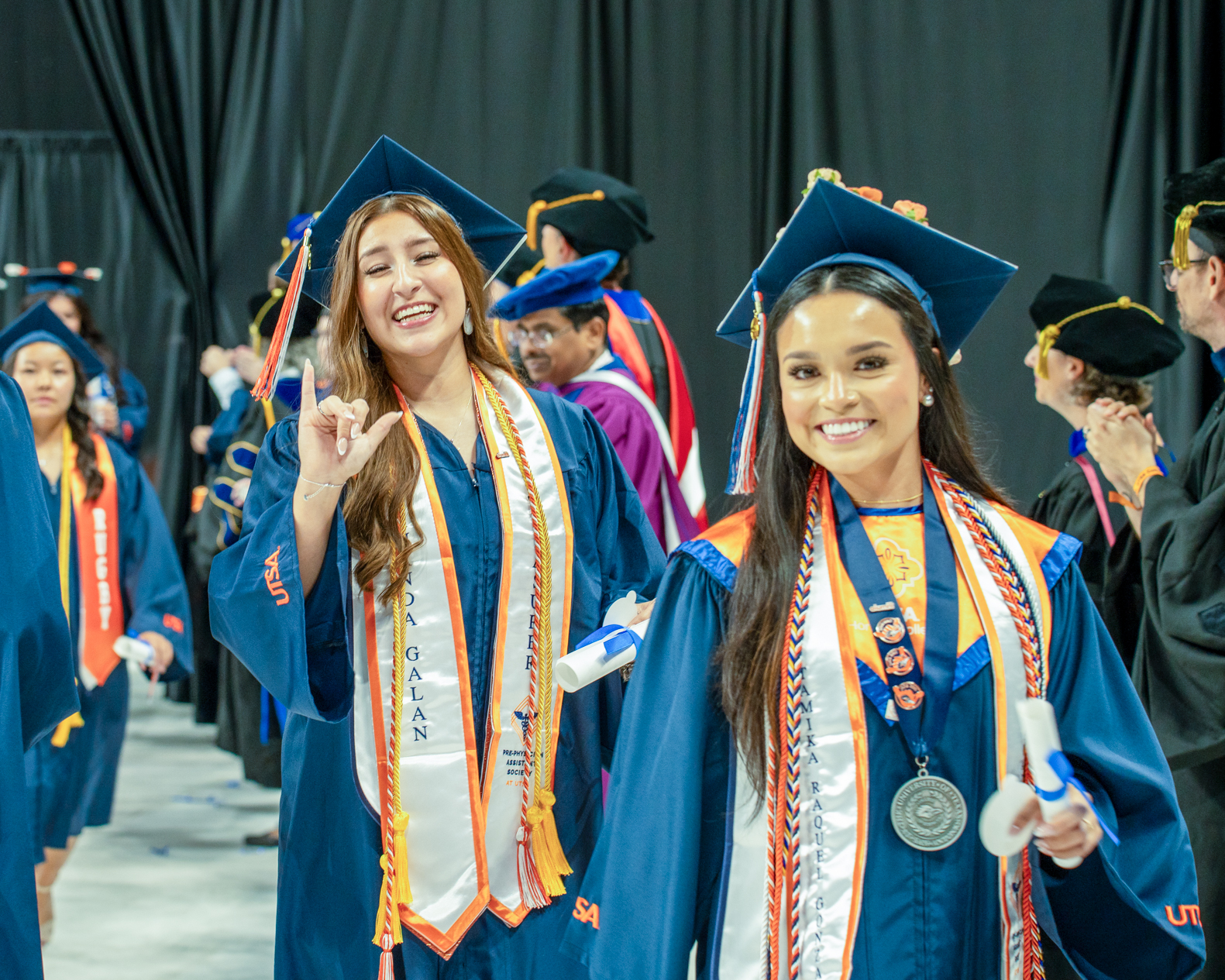 Two women in cap and gown during graduation one making the birds up UTSA gesture
