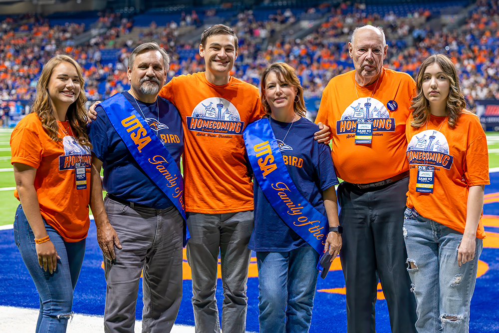 A family of 6 people at standing in a football stadium, 4 are wearing orange Homecoming teeshirts and 2 are wearing blue sashes that say Family of the Year.