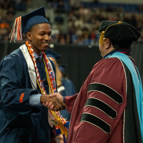 Two students in caps and gowns standing with their arms extended in celebration amid graduating students in the Alamodome
